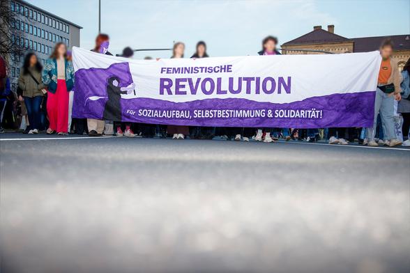 Im unteren Bereich des Bildes ist der Teer einer Straße zu sehen, welcher mit einem Blitz augehellt wird. 
In der oberen Hälfte ist ein Transparent mit dem Text "Feministische Revolution - Für Sozialaufbau, Selbstbestimmung & Solidarität", welches von Menschen getragen wird zu sehen