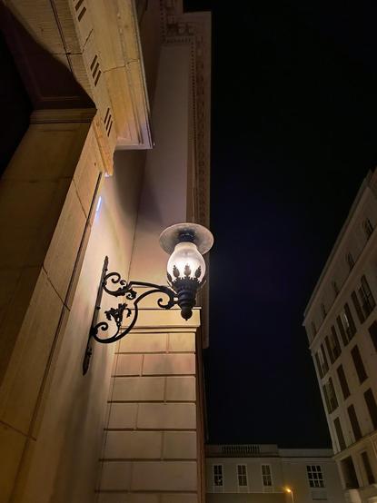 Links im Bild ist eine schöne altmodisch verzierte Laterne aus gusseisernem schwarzen Metall und gewölbten Glas an eine beigefarbene Hauswand eines alten, großen, massiven Hauses im Schinkelschen Stil montiert und erleuchtet die Wand. Im Hintergrund und rechts befinden sich weitere, ähnliche Häuser. Der Himmel oberhalb ist schwarz wie die Nacht.