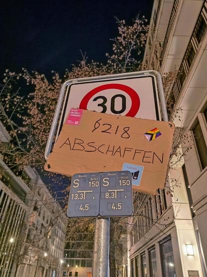In einer Straße mit großen, hohen, Gebäuden in beige unter einer nigen jungen Bäumen steht unter einem schwarzen Nachthimmel ein Verkehrsschild mit Tempo 30 und zwei kleineren blauen Schildern zum Grundwasserzugang. Dazwischen wurde ein Demoschild aus hellbrauner Pappe gesteckt, auf dem steht: Paragraph 218 abschaffen.