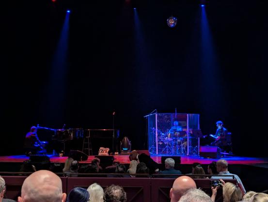 A theater stage lit in soft red and purple. A grand piano and drum set are being set up.