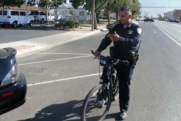 A City of Soledad, California police officer on a bicycle has paused to explain something to someone, as his hand gesture indicates.