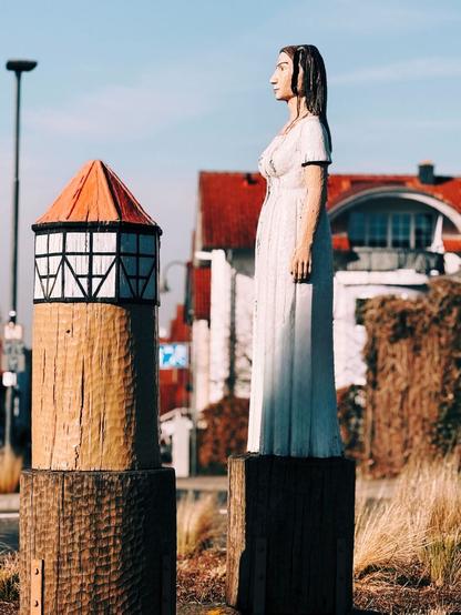 Zwei Holzskulpturen auf einem Kreisverkehr in Burg Gräfenrode, bekannt als „Liesel-Kreisel". Die linke Skulptur stellt den Liesel-Turm dar, mit einem rot gedeckten Dach und einer Fachwerkstruktur. Rechts steht die Figur der Liesel, eine Frau in einem weißen Kleid mit langen dunklen Haaren, die ruhig in die Ferne blickt. Beide Skulpturen stehen auf massiven Holzsockeln. Im Hintergrund sind Gebäude mit roten Dächern, eine Straßenlaterne und trockene Gräser zu sehen. Die warme Sonneneinstrahlung betont die Holzstruktur und verleiht der Szene eine stimmungsvolle Atmosphäre.