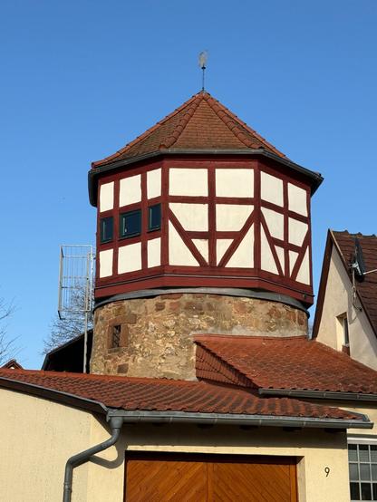 Nahaufnahme des Liesel-Turms, eines historischen Wehr- oder Gefängnisturms mit rundem Sandsteinmauerwerk und einem achteckigen Fachwerkaufsatz in Rot-Weiß.
Der Turm hat ein spitz zulaufendes Dach mit einem Wetterhahn an der Spitze. Im Hintergrund ist ein klarer blauer Himmel zu sehen.