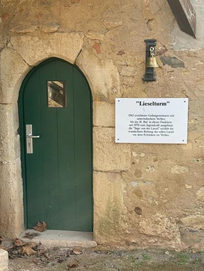 Detailaufnahme der grünen Holztür des Liesel-Turms mit einem kleinen Fenster. Daneben hängt eine bronzefarbene Glocke an der Wand. Eine weiße Informationstafel beschreibt die Geschichte des Turms, der 1563 als Gefängnisturm mit Verlies errichtet und später zum Jugendcafé umgebaut wurde.
Am Boden liegen einige trockene Blätter.