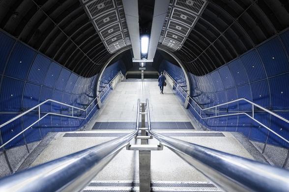 Look-down shot with a wide-angle lens of stairs going down to underground platforms through a dark blue, black and grey tube with a Sci-Fi look. The silhouette of a man with a dark blue suit appears on the right at the bottom of the first set of stairs.
