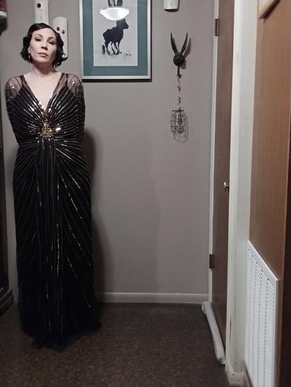 A lone woman at the left edge of the picture is standing in a floor-length black gown with gold sequins and beads. Her hair and makeup is inspired by 1920s styles.