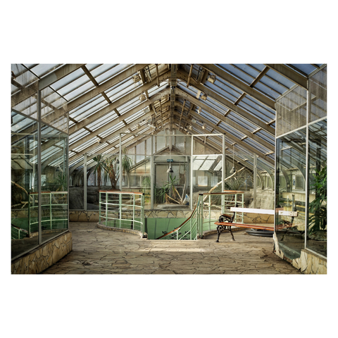 This is a wide angle landscape format colour image of the interior of a disused wing of a glasshouse at Budapest Zoo. The image is a confluence of vertical and horizontal lines and resulting angles, giving it a geometric feel. The pitched glass ceiling lets light through to the shabby vintage interior; the colours soften the overall geometric effect. Unused animal enclosures sit to either side of the frame, and in the centre is the top of a spiral staircase. To the right of the top of the stairs is an old wooden bench - the worn wood gleams in the sunlight. Taken with a Sony RX100.