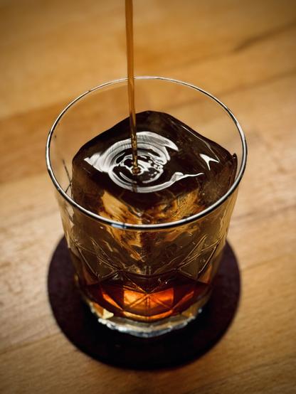 dark brown cocktail being poured over a large rock of clear ice into a glass with a deep maroon coaster underneath it
