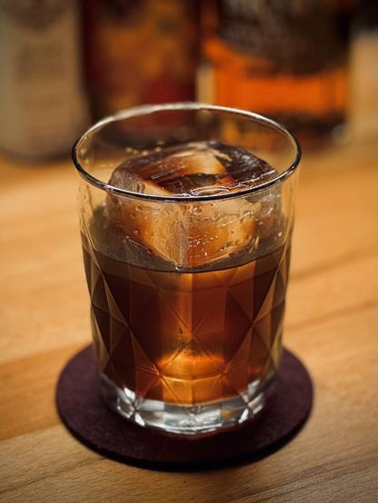 dark brown cocktail with a large rock of clear ice in a faceted glass with silver rim with bottles of Fee Brothers black walnut bitters, Northern Monk Chocolate-dunked Digestive Stout, and Sazerac rye whiskey in the background