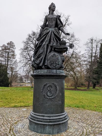 A full shot of a bronze statue depicting a young Catherine the Great standing on a round plinth in a park setting. She wears a long, detailed dress and a small crown, with her right arm extended. To her right, on a smaller stand, is a crown and a key. The plinth is decorated with a wreath and has the inscription 'Sophie Auguste Friederike', her birth name. The statue is surrounded by a circular cobblestone path, with green lawn and bare trees visible in the background.