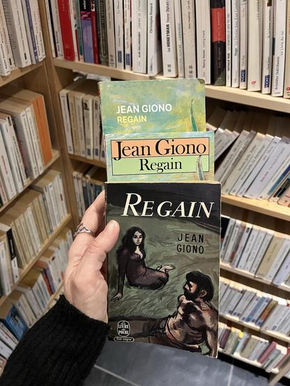 3 exemplaires de livres du même titre et du même auteur. Il s’agit de « Regain » de Jean Giono. Je tiens les 3 livres dans ma main gauche pour prendre la photo. En arrière plan, une bibliothèque remplie de bouquins.