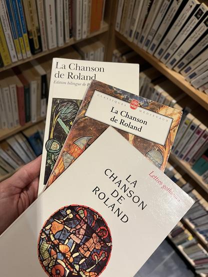 3 exemplaires de livres du même titre et du même auteur. Il s’agit de « La Chanson de Roland ». Je tiens les 3 livres dans ma main gauche en éventail pour prendre la photo. En arrière plan, une bibliothèque remplie de bouquins.