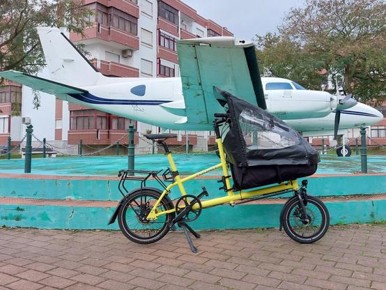 Uma bicicleta se carga amarela, rodas baixas, frente de carga com capota preta em frente a um pequeno avião branco num jardim