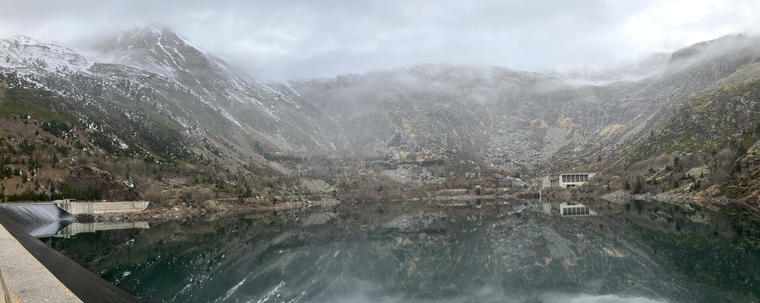 Vista panoràmica del pantà de Sallente amb les instal·lacions de la hidroelèctrica al fons i les muntanyes que envolten el pantà, quedant tot el conjunt reflectit sobre l'aigua plana com un plat del embassament.