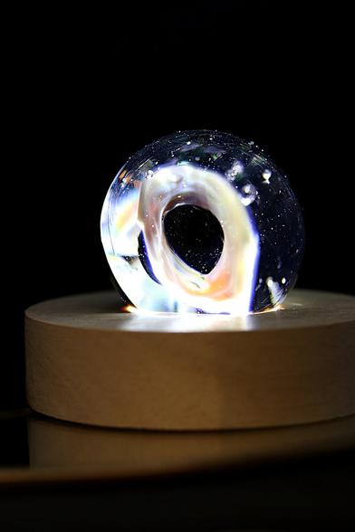 Photographie d'une sculpture en verre en forme d'anneau, évoquant un trou noir lumineux.   L'œuvre, aux reflets irisés et ponctuée de fines bulles d'air, repose sur un socle en bois clair.   Un éclairage bien placé traverse le verre, créant une aura fascinante et une illusion de profondeur infinie.   L'arrière-plan sombre accentue l'effet cosmique et mystérieux de la pièce.