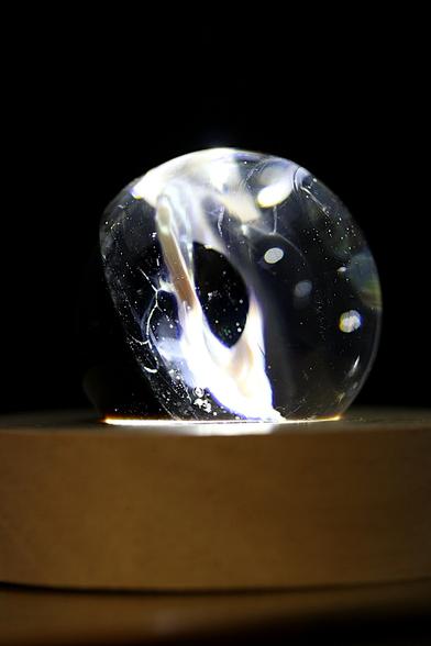 Photographie d'une sculpture en verre en forme d'anneau, évoquant un trou noir lumineux.   L'œuvre, aux reflets irisés et ponctuée de fines bulles d'air, repose sur un socle en bois clair.   Un éclairage bien placé traverse le verre, créant une aura fascinante et une illusion de profondeur infinie.   L'arrière-plan sombre accentue l'effet cosmique et mystérieux de la pièce.
