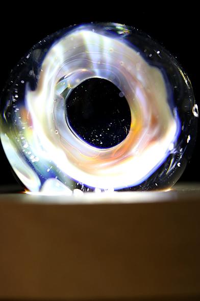 Photographie d'une sculpture en verre en forme d'anneau, évoquant un trou noir lumineux.   L'œuvre, aux reflets irisés et ponctuée de fines bulles d'air, repose sur un socle en bois clair.   Un éclairage bien placé traverse le verre, créant une aura fascinante et une illusion de profondeur infinie.   L'arrière-plan sombre accentue l'effet cosmique et mystérieux de la pièce.