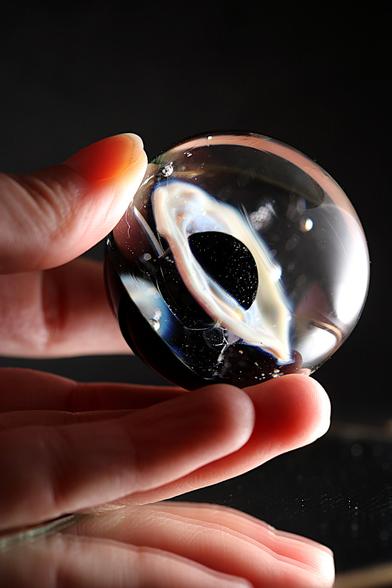 Photographie d'une sculpture en verre en forme de sphère, tenue délicatement entre les doigts d'une main. L'œuvre présente un trou noir central entouré d'un anneau lumineux aux reflets irisés, évoquant une singularité cosmique. De fines bulles d'air sont emprisonnées dans le verre, ajoutant de la profondeur à la pièce. L'éclairage accentue la transparence et les jeux de lumière, contrastant avec l'arrière-plan sombre qui met en valeur la finesse et la délicatesse de cette création."