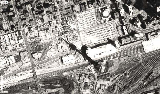 Aerial imagery showing the CN Tower casting a shadow on surrounding land in 1985. At the time, much of the nearby area were parking lots, warehouses, and rail yards.