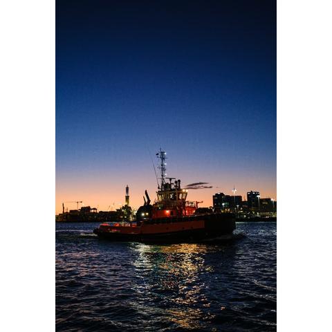 A tugboat returns to the port of Genova at the sunset. The tugboat is painted in orange. The lighthouse is in the background.