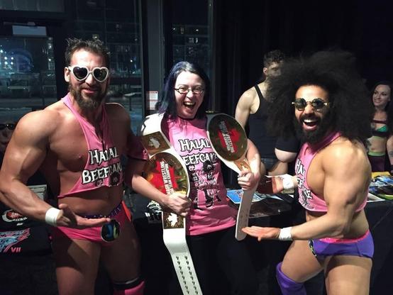 A group of three individuals poses together, wearing matching pink tank tops with "Halal Beefcake" printed on them. Two very muscular men stand on either side of a woman, all smiling enthusiastically. The woman holds two championship belts.