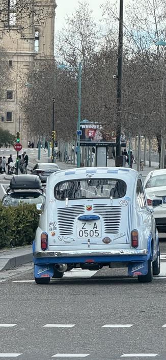 Un Seat 600 clásico, azul con franjas blancas, circula por una calle urbana. Su matrícula “Z-A 0505” indica registro en Zaragoza. La parte trasera muestra adhesivos, incluido el logo de Abarth y una pegatina conmemorativa de los 60 años del modelo. Tiene modificaciones como faldones con “SEAT 600 E” y un escape grande. En el fondo, hay semáforos, una parada de bus con publicidad, peatones y árboles sin hojas, sugiriendo invierno. A su izquierda, un coche oscuro aparcado en la acera; a la derecha, otro vehículo circulando.