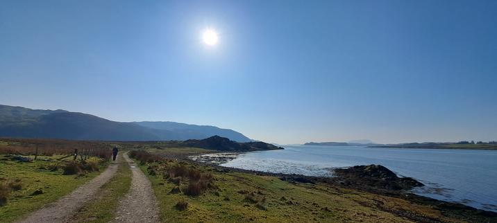 On the eastern bank of Seil Sound, a solitary person walks along a track. Grassland runs gently to the shore; in the background are the higher hills of the Degnish peninsula. Along the Sound is the island of Torsa, where the Cuan Sound leads to Easdale Bay.  In the hazy far distance is Scarba. The sun shines down from a clear blue sky.