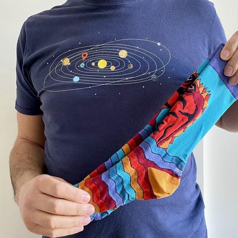A person is holding a colorful sock with a zigzag pattern and a cartoonish red creature on it. They are wearing a navy blue t-shirt featuring a graphic of the solar system with planets and a location pin.