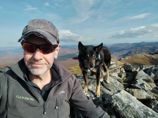 Selfie, myself and Moray the dog, we're at the top of a mountain, it is rocky and grassy. Blue sky with clouds.