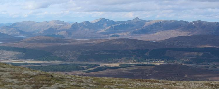 A hazy view of a mountain range in the middle distance. Mostly grassy in the foreground with some rocky bits. The distant hills have very few snow patches on them. Cloudy, blue sky.