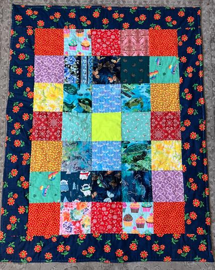 A colorful quilt made from 35 squares arranged in a 7x5 rectangle; all of the fabrics are prints, except the center fabric, which is a solid neon yellow; the border is a navy vintage print with orange flowers.