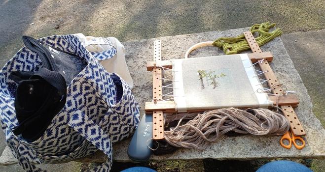 A partvworked stitched artwork, on a wooden frame, lies upon a stone table. Green and brown wool sits next to it. A bag and spectacle case sits to the left. The scene is lit by dappled sunlight