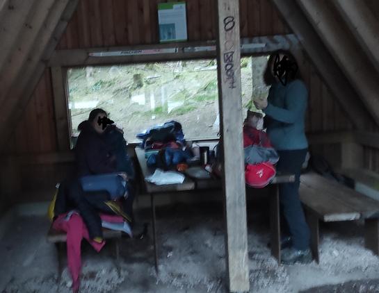 2 Frauen in einer Schutzhütte mit Tisch und 2 Bänken und rückwärtigen Fenster. Auf dem Tisch stehen Kocher, Tassen, Teller und Frühstückszeug