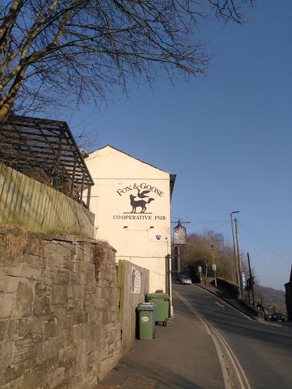Photo of a white building (the pub) by a road with a big painting of the Fox and Goose logo on it.