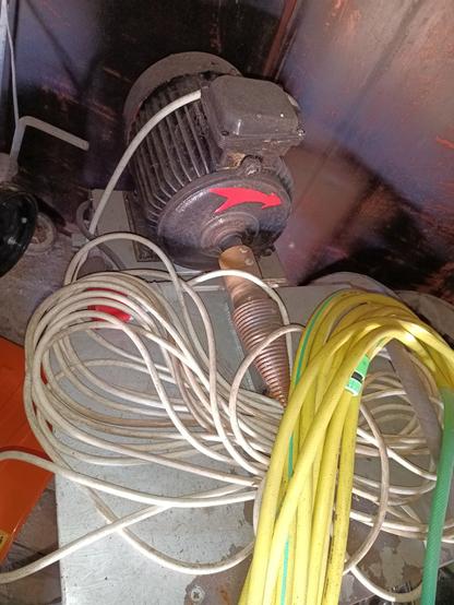 A bad photo of a large three phase electric motor with a 70mm diameter threaded cone mounted directly on the shaft. A large red arrow indicates it turns clockwise looking from the front. It's mounted on a table. A coiled up three-phase power cable is on it, and a yellow garden hose on top of that.