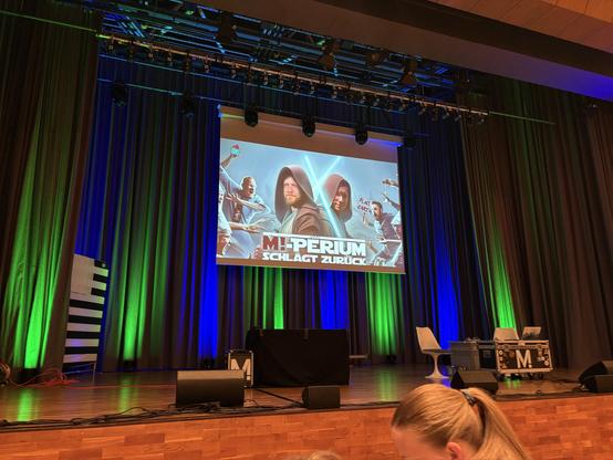 Ein Foto von einer Bühne in einem Auditorium. Blaue und grüne Vorhänge hängen hinter der Bühne. Ein Bildschirm zeigt ein Bild mit dem Text "M-Perium schlägt zurück". Auf dem Bild sind zwei Personen in Jedi-ähnlichen Gewändern zu sehen. Vor der Bühne stehen Lautsprecher und andere Bühnenelemente. Im Vordergrund ist der Hinterkopf einer Person zu sehen. Die Bühne ist aus Holz.

Bereitgestellt von @altbot, generiert mit Gemini