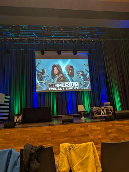 Leere Bühne im Hintergrund eine Leinwand das an Star wars erinnert und auf dem steht: "Das M!-mperium schlägt zurück." Und Reini und Nicolas als Jedi Ritter mit Lichtschwerter gezeigt werden.