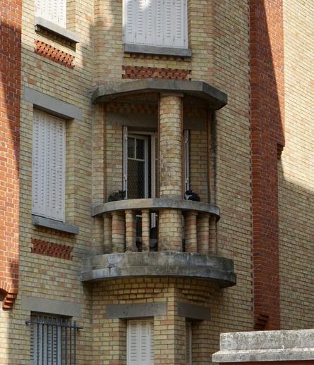 Photo d’un balcon en quart de cercle sur une façade en brique jaune rehaussée de quelques touches de brique rouge. Les montants du garde corps du balcon sont eux-mêmes constitués d’empilements de briques arrondies. Un pilier central en brique jaune un peu plus épais vient relier le dessus et le dessous du balcon, qui sont en béton. Ce balcon a l’air vraiment tout petit. Deux chats sont installés sur sa rambarde.