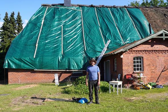 Mann mit Sonnenhut steht vor Haus mit grüner Plane auf dem Dach