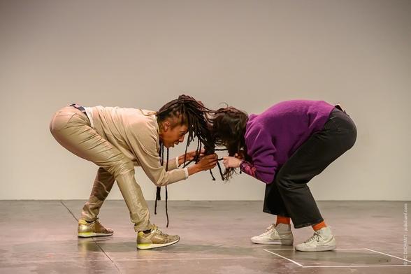 Angele-Rohart & Marie-Julie Sylvette – Hair Together
Deux femmes sont penchées l’une vers l’autre, leurs têtes presque collées, comme reliées par leurs cheveux emmêlés. Elles adoptent une posture chorégraphiée, l’une vêtue d’un pantalon doré et l’autre d’un pull violet. L’arrière-plan est un mur blanc sobre.