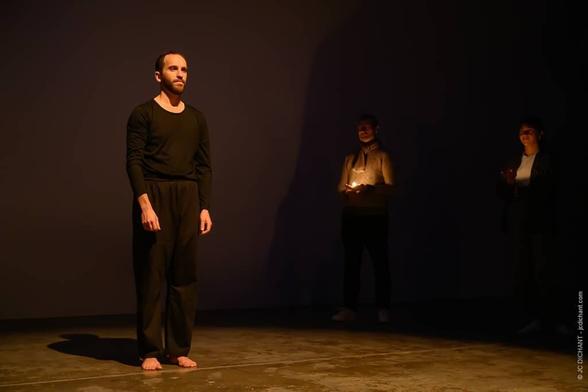 Brieuc Guinard – Break the Cycle (Photo 2)
Un homme en tenue noire se tient debout sur scène, éclairé par une lumière douce et chaude. En arrière-plan, deux autres personnes tiennent des bougies, leurs visages à peine visibles dans l’ombre.
