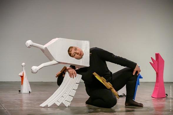 Charlotte Larouche – La File (Photo 1)
Une femme vêtue de noir est agenouillée sur scène avec un grand accessoire en carton blanc en forme de bâtiment sur la tête. Elle semble en équilibre instable, tenant des formes colorées dans ses mains. En arrière-plan, d’autres structures en carton rappellent des éléments architecturaux.