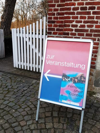 Aufsteller "Zur Veranstaltung. Feminist Shelf Control", dahinter ein weißes Holztor, Backsteinmauern des Gebäudes