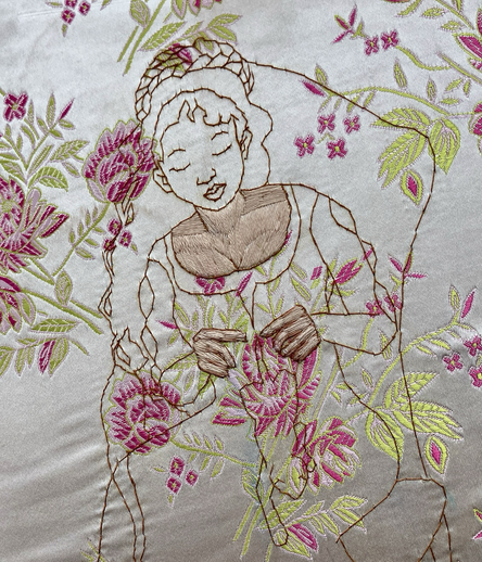 An outline of a woman knitting, stitched in golden brown thread on a cream brocade with pink roses and green leaves, with her hands and chest filled in a pale peachish tone.