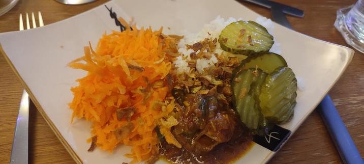 A square plate of food with a bit pike or bright orange carrots that have been grated and soaked in lemon juice with a bit of sugar, a mound of white rice, a big spoonful of the babi ketjap, sprinkled with crispy fried onions, and some pickles on the the side.