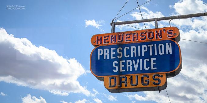 The blue and orange neon sign saying "HENDERSON'S PRESCRIPTION SERVICE DRUGS" with blue sky and clouds in the background.