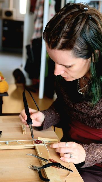 English A woman with partly forest-green hair works at her desk with a soldering iron on small glass pieces containing pressed autumn leaves.
Français Une femme aux cheveux en partie vert forêt travaille à son bureau avec un fer à souder sur de petites pièces en verre contenant des feuilles d'automne pressées.