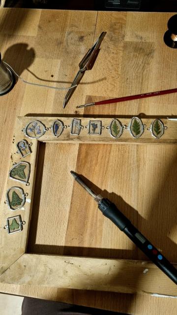 English The table of a craftswoman's workshop with glass pieces containing pressed flowers and leaves with silver outlines and soldered rings on both sides. Work tools can be seen: soldering iron, brush, etc.
Français La table d'un atelier d'artisane avec des pièces en verre contenant des fleurs et feuilles pressées aux contours argentés et avec des anneaux soudés de part et d'autre. On aperçoit des outils de travail : fer à souder, pinceau etc.