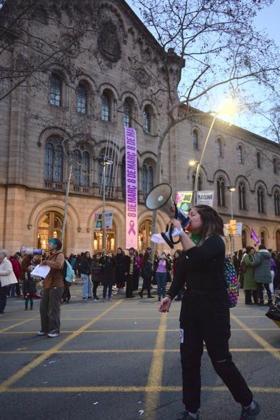 The girl stands out at the centre, wearing all-black attire and carrying a colourful backpack, while addressing the crowd through a megaphone held firmly in her hand. In front of her, people hold signs bearing messages of solidarity. In the background, the tan stone building (Universitat de Barcelona historic building) features a prominent poster with purple ribbon, reading "8 MARÇ," which translates to March 8th.

La noia surt destacada al centre, vestint tota la roba negra i portant una bossa multicolor, mentre dirigeix la gent a través d'un megàfon que té ben agafat a la mà. Davant seu, les persones tenen senyals amb missatges de solidaritat.  Al fons es veu l'edifici de pedra fosca (l'edifici històric de la Universitat de Barcelona) i un cartell molt visible amb una cinta violeta amb el lema "8 MARÇ".