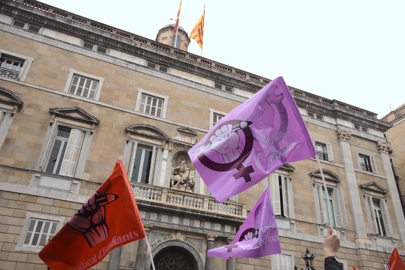 The image shows people holding two purple flags bearing the feminist symbol and red flags, in front of the Palace of Catalonia Government. Atop the building is the flag of Catalonia. The building itself features a prominent archway above the entrance, with a small balcony.

La imatge mostra gent que porta dues banderes de color lila amb el símbol feminista i banderes roges davant del Palau de la Generalitat. Al damunt hi ha la bandera de Catalunya. El mateix edifici presenta una arcada prominent sobre l'entrada, amb un petit balcó.